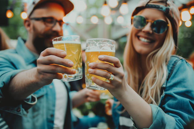 Ein Mann und eine Frau stoßen bei einer Outdoor-Party mit Bier in hochwertigen Nachtmann Gläsern an. Beide tragen lässige Kleidung und Sonnenbrillen, während sie fröhlich in die Kamera lächeln.