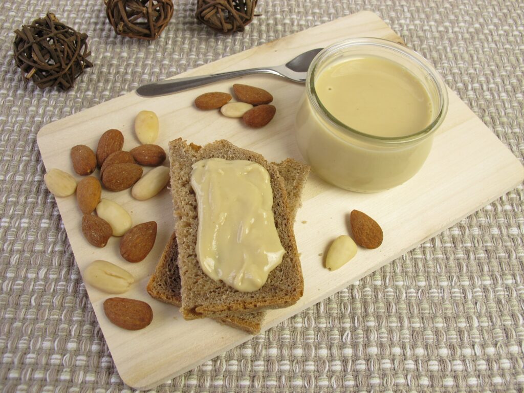 Ein Glas mit cremigem Nussmus neben zwei Scheiben Brot mit Nussmus und einer Handvoll Mandeln auf einem Holzbrett.