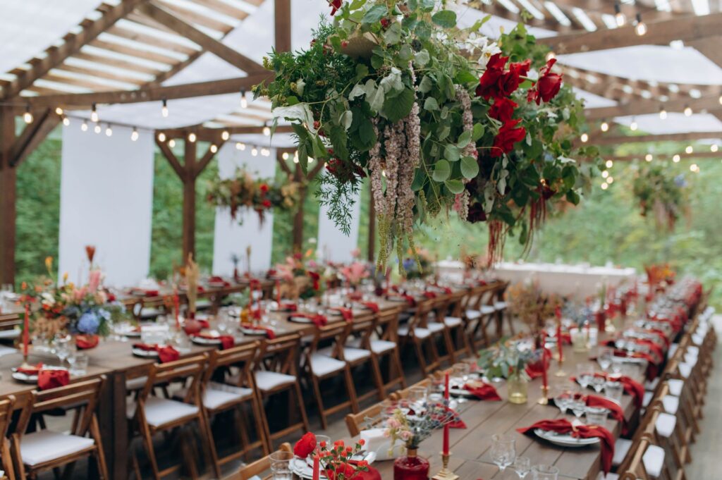 Eine festlich gedeckte lange Tafel unter einem überdachten Holzpavillon im Freien, dekoriert mit üppigen hängenden Blumenarrangements, roten Stoffservietten und Kerzen, die eine elegante und romantische Atmosphäre für ein Outdoor-Event schaffen