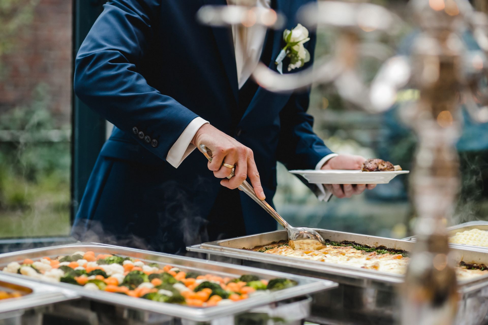 Ein eleganter Mann im Anzug serviert sich Essen von einem hochwertigen Buffet mit dampfenden Speisen, darunter buntes Gemüse und ein Gratin, während er eine festliche Veranstaltung besucht.