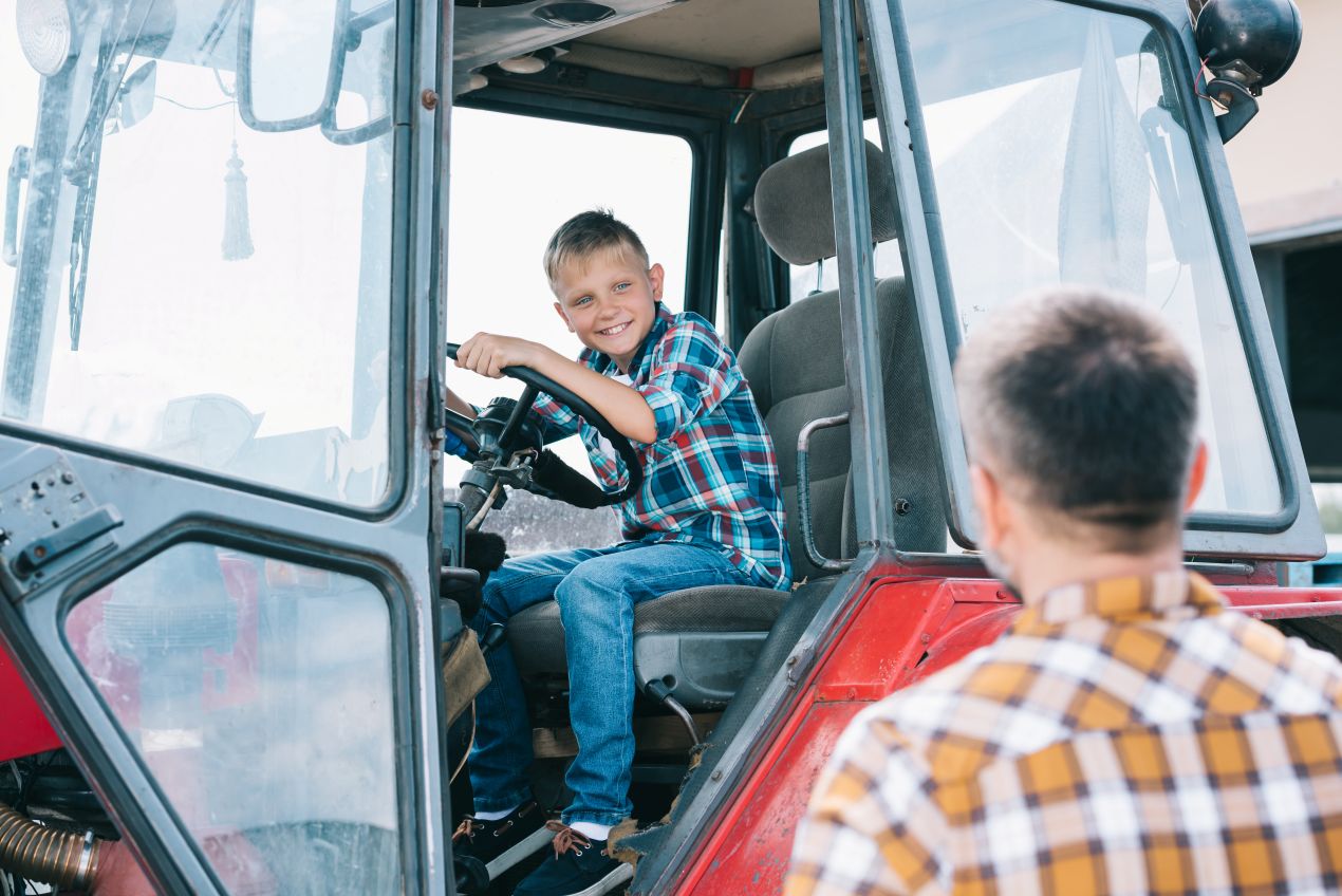 Junge sitzt lachend am Steuer eines Traktors auf einem Erlebnisbauernhof NRW, während ein Erwachsener ihn anleitet.